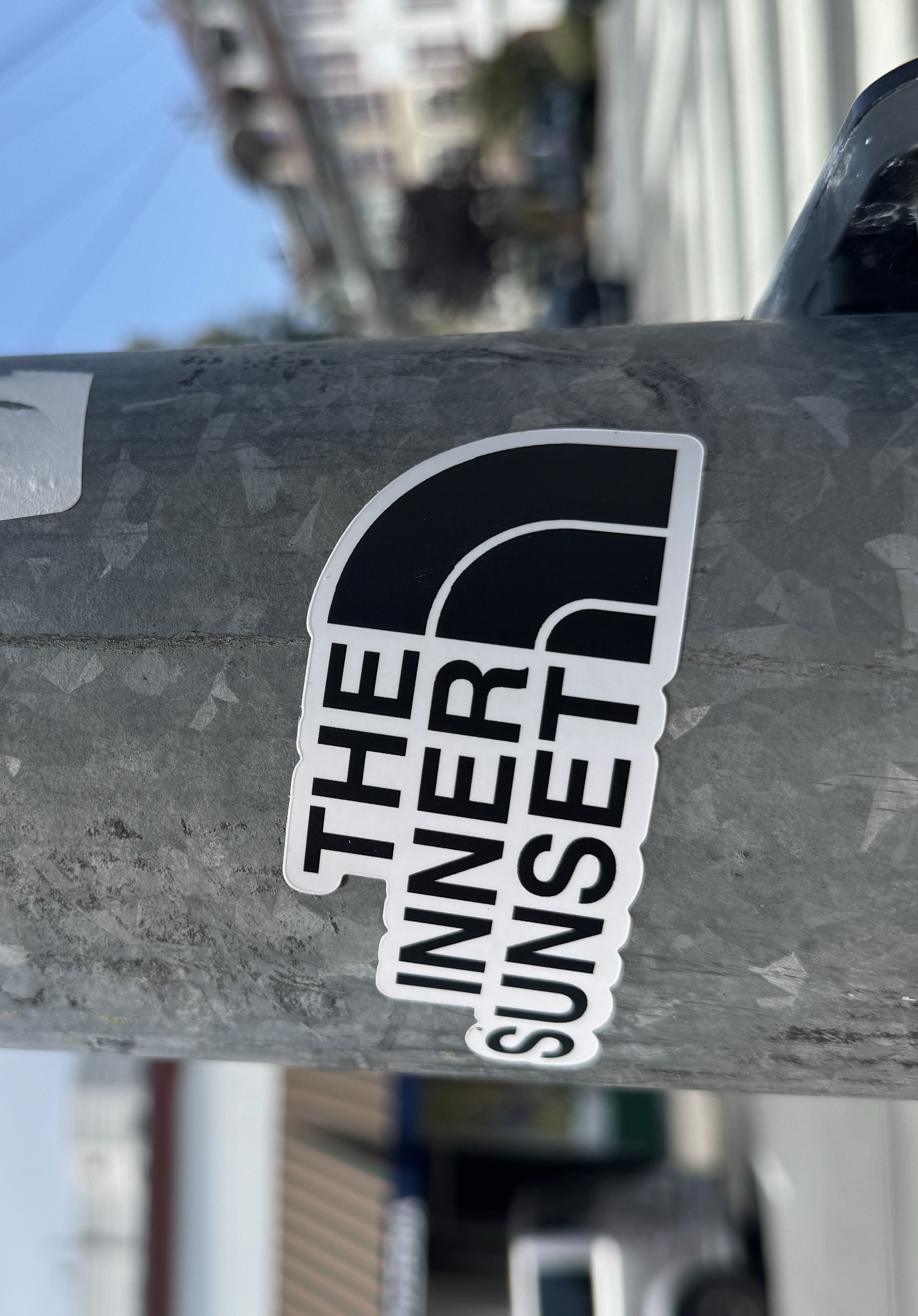 A close-up photo of the surface of a lamppost on a street corner, with a sticker on it, in the middle, styled like The North Face logo but instead reading 'The Inner Sunset'.